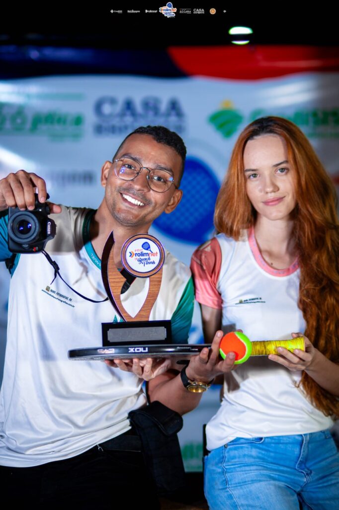 Dupla de fotógrafos sorrindo, com uma câmera, um troféu de torneio e uma raquete com bola, diante de painel de patrocinadores.
