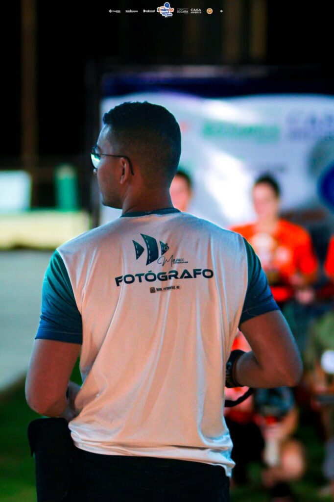Fotógrafo de costas, com camiseta escrita FOTÓGRAFO, em evento noturno com pessoas desfocadas ao fundo.