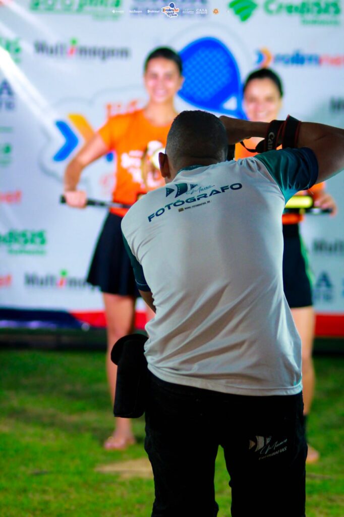 Fotógrafo de costas, com camiseta escrita FOTÓGRAFO, fotografando participantes diante de um painel de patrocinadores.