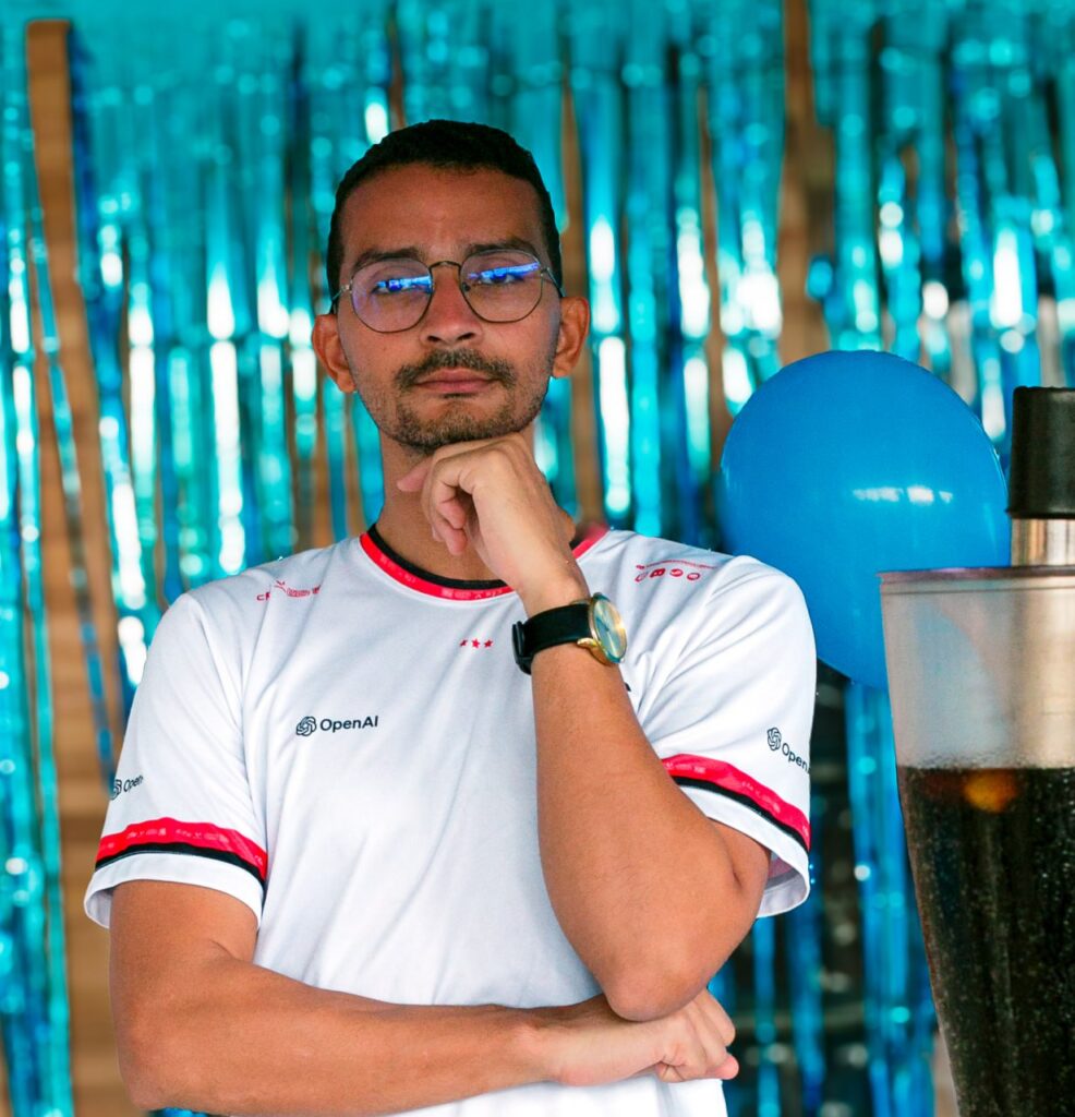 Retrato de homem de óculos com a mão no queixo, diante de uma cortina metálica azul e um balão.