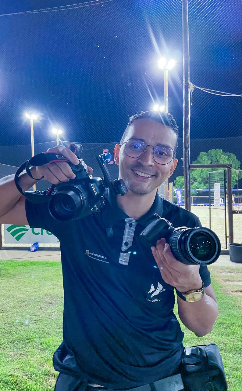 Fotógrafo sorrindo com duas câmeras profissionais em uma quadra iluminada por refletores, à noite.